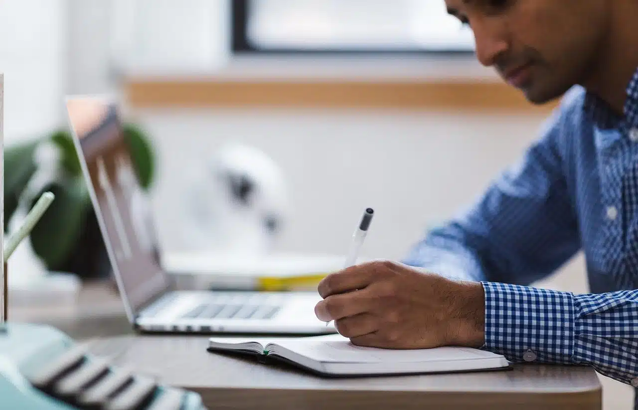 Man writing things down in his notebook, next to his laptop.