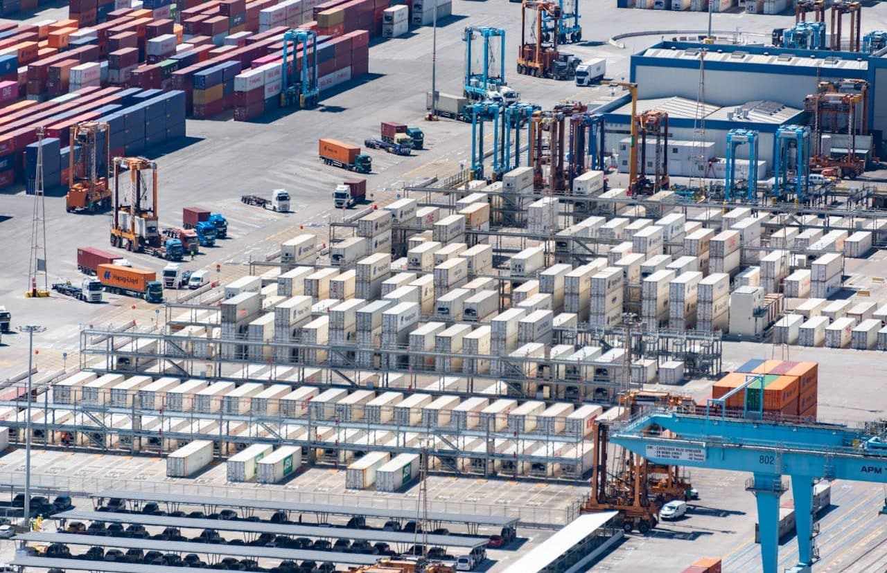 Distant view of cargo containers in a port, with trucks and cranes.